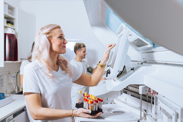 A laboratory technician is organizing blood samples while a colleague works on equipment in a bright lab.