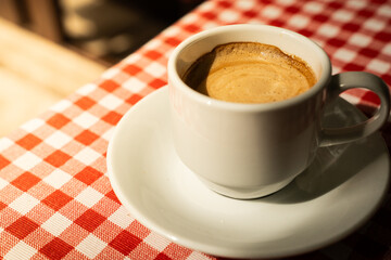Cup of coffee on red checkered tablecloth. White cup with hot coffee on table in cafe with red checkered tablecloth. Concept of breakfast, energy drink, lifestyle and cafe culture.