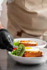 Chef plating gourmet toast with sunny-side-up egg, fresh greens, and cherry tomatoes in a professional kitchen. Culinary presentation, food styling, and restaurant service concept.