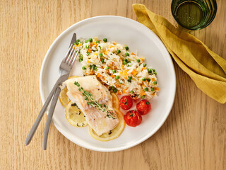 A plate features a portion of cooked cod fish beside vibrant rice mixed with peas, carrots, and cherry tomatoes. This nutritious dish is perfect for lunch or dinner