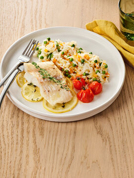 A flavorful plate features tender cod fish garnished with lemon slices, served alongside colorful rice mixed with peas and carrots, and cherry tomatoes