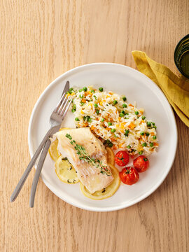 A plate features tender cod fish alongside fluffy rice mixed with green peas and carrots, garnished with lemon slices and fresh tomatoes. A simple yet appealing meal