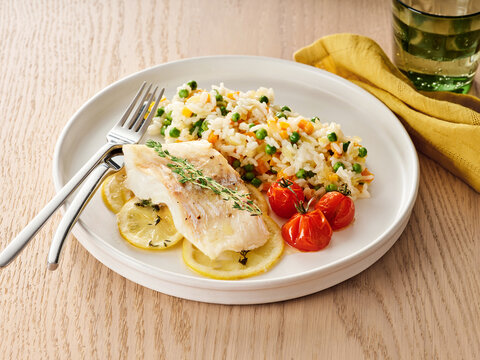A delicious meal features cod fish alongside fluffy rice mixed with colorful vegetables. The dish is artistically presented with lemon and cherry tomatoes, inviting and fresh