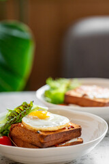 Close-up of brunch with sunny-side-up egg on toast, fresh greens, and cherry tomato in a white bowl. Natural light, cozy indoor setting with plant in background.