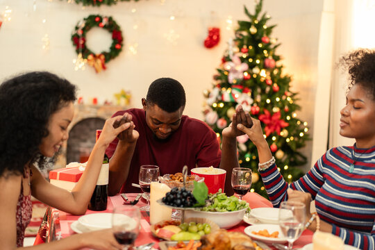Diverse Black family holding hands, bowing heads. Praying at Christmas dinner table. Festive meal, holiday celebration. Christmas tree and decorations background. Seasonal atmosphere - Powered by Adobe
