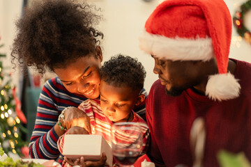 A joyful African American family support their son to open a glowing gift box on Christmas season at dinner table. Moment of magic, love, and surprise on holiday party. Love and bonding people