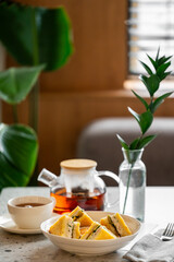 Breakfast setting with triangular sandwiches, tea in cup and teapot, green plant decor, cozy natural light, minimalistic tableware, inviting and clean composition.