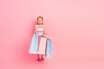 Adorable young girl holding shopping bags on pink background, looking amazed, expressing excitement and charm