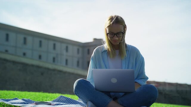 Blonde woman with glasses sitting on grass  city park, using a laptop for remote work or online education, enjoy sunny day while working as digital nomad.Business, freelance concept. Lifestyle