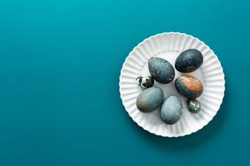 Decorative Easter eggs with various textures and colors are arranged neatly on a white plate, resting on a vibrant teal surface, creating a festive spring atmosphere