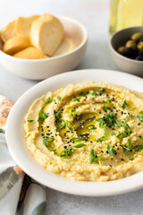 A bowl of creamy hummus topped with olive oil and herbs sits beside fresh bread and a bowl of olives. The light setting showcases a simple, inviting meal