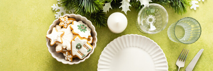 A bright green Christmas table setting with an empty plate and cutlery. The table is decorated with spruse branches and white tree ornaments, creating a festive vibe, banner