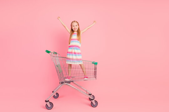 Cheerful young girl in a colorful dress standing in a shopping cart with raised arms against a pink background - Powered by Adobe