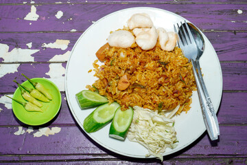 fried rice dish on a white plate with toppings of sliced ​​cabbage, cucumber and some crackers isolated on a purple wooden table surface.