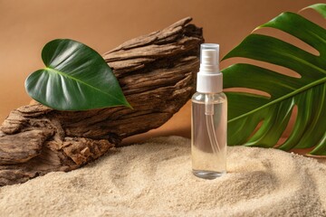 A clear spray bottle with liquid sits on sand next to driftwood and tropical leaves