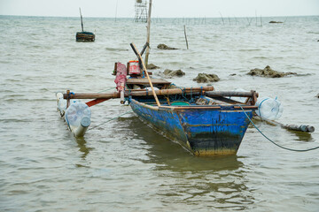 view of a Javanese man with shoulder-length hair drinking through a straw from a glass outdoors with a cafe in the background with wooden chairs and tables and palm plants.