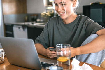  Gen Z woman working from home at cozy kitchen table with laptop and phone. Digital nomad...