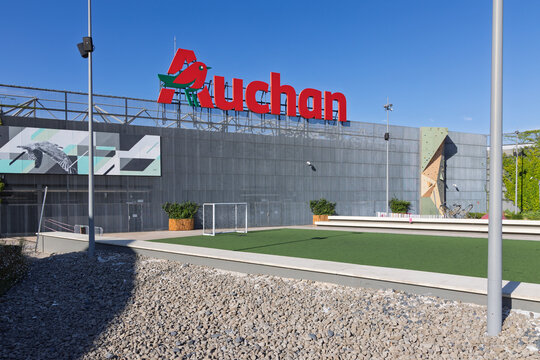 The Auchan hypermarket logo is prominently displayed on the dark, modern exterior of a large store. The foreground features a soccer pitch and gravel landscaping. Setubal, Portugal. 18 August 2025.