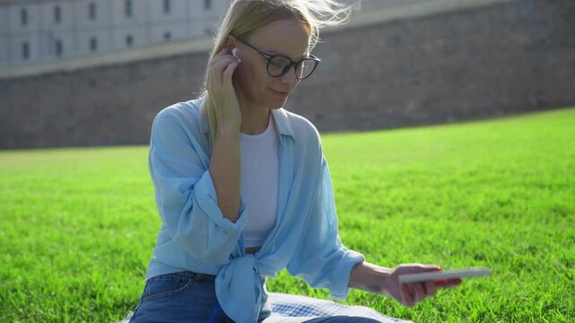 Happy young woman putting in wireless earbuds and choosing song on smartphone while sitting on green lawn in public park on sunny day, enjoying her free time outdoors