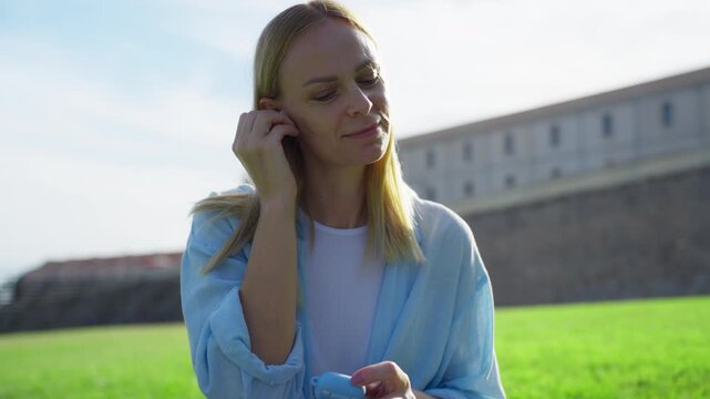 Young woman relaxes on park grass, takes wireless earbuds from their case, taps smartphone to pick song and smiles as enjoy music on sunny summer day