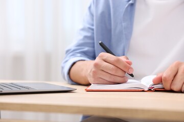 Man taking notes while passing online test via laptop at wooden table indoors, closeup