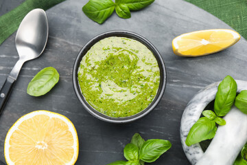 Tasty green pesto in bowl, basil leaves and lemon on black table, top view