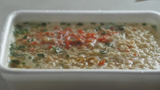 Close-up of appetizing broth with vermicelli instant noodles brewing in a plastic container with boiling water, covered with a plastic lid. Hot instant noodles, quick homemade meal