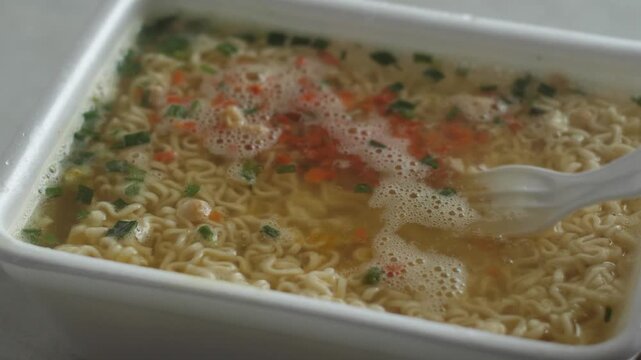 Close-up of appetizing broth with vermicelli instant noodles brewing in a plastic container with boiling water, covered with a plastic lid. Hot instant noodles, quick homemade meal