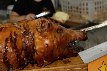 A whole roasted pig at a lively outdoor food event. A food vendor prepared a sandwich in the background, highlighting a bustling atmosphere filled with joy and celebration