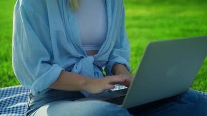 Young woman, not easily identifiable, work on laptop while seated on picnic blanket in grassy field, fully engaged in remote work and digital nomad lifestyle.Freelance business concept