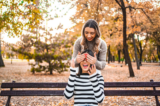 One woman surprises another sitting on a bench by covering her face with her palms