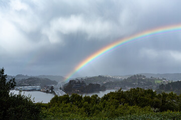 A rainbow arcs across the sky over a landscape featuring a bridge with traffic, water, and green hills under a cloudy sky.