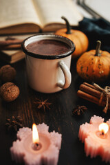 Cozy autumn scene with mug of hot chocolate, book, pumpkins, spices and candles on rustic dark wooden background, warm seasonal atmosphere