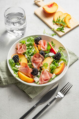 Fresh salad with peaches, arugula, feta, prosciutto and berries in a plate on a light background