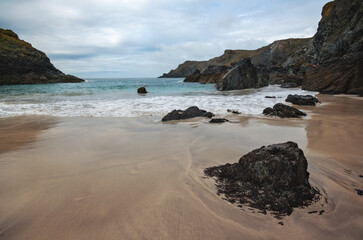 Large dark rocks on smooth sand beach at Kynance Cove, ocean in background