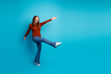 Obraz premium Joyful young woman wearing a brown sweater and jeans, posing with a kick motion against a blue background