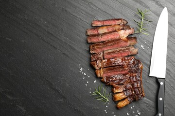 Pieces of delicious grilled beef steak with rosemary, salt and knife on grey textured table, flat...
