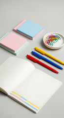 Angled shot of an open notebook with blank pages, stacks of pink and blue sticky notes, a dish of colorful paperclips, and pens in red, blue, and yellow on a gray surface, showcasing a creative worksp