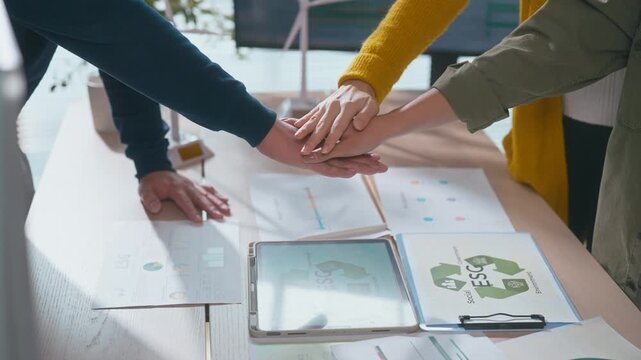 Asian business team discusses ESG strategy in office with documents on table, then stack hands to show unity, teamwork, and commitment to sustainability goals.