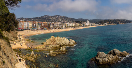 Lloret de Mar's main beach, with coastal buildings and mountains in the background. The partly cloudy sky contrasts with the blue sea and golden sand. Spain