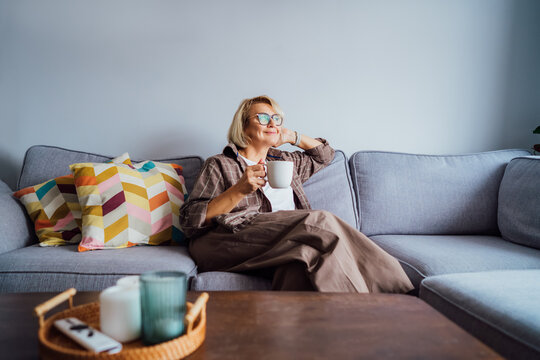 Middle aged smiling woman sitting on sofa and looking away while drinking coffee or tea. Mature senior woman relaxing after housekeeping, home cleaning. Portrait of relaxed female resting at home.