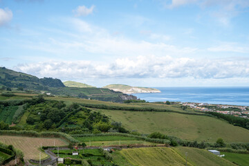Naklejka premium Azores landscape with panoramic views of green hills, pastures sloping down to the coast, a coastal village and the Atlantic Ocean stretching to the horizon