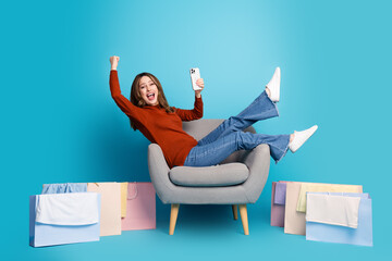 Excited young woman holding phone seated comfortably amid colorful shopping bags on a bright blue background