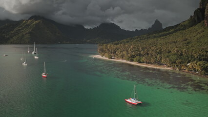 Stunning aerial view of yachts in turquoise lagoon bay, tropical island beach in French Polynesia, green mountains and dramatic cloudy sky. Remote wild nature paradise, exotic summer travel background