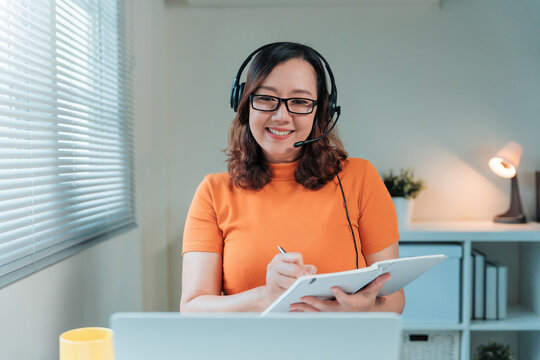 Woman wearing headset making notes, looking at camera and smiling, working from home office, doing online learning or customer service