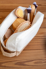 A wooden comb, a wooden roller for restorative facial massage, lymphatic drainage, and wrinkle reduction, and bamboo makeup brushes in a natural linen bag on a wooden background. Zero-waste self-care