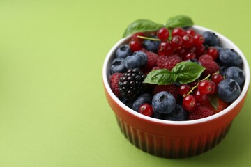 Different ripe berries and basil leaves in bowl on green background, closeup. Space for text