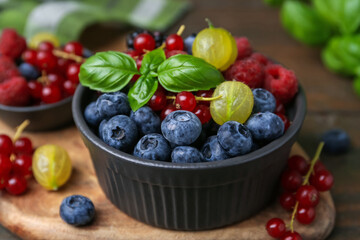 Different ripe juicy berries and basil in bowl on wooden table, closeup