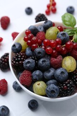 Different ripe juicy berries and basil in bowl on white tiled table, closeup