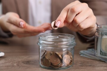 Woman putting coin into glass jar at wooden table indoors, closeup
